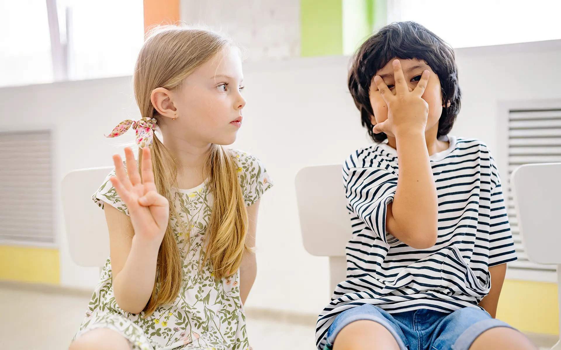 Two children sitting indoors, engaging in a playful hand gesture activity. The girl on the left, wearing a floral dress with a ribbon in her hair, holds up four fingers while looking at the boy. The boy on the right, dressed in a striped shirt and denim shorts, makes a playful gesture by spreading his fingers over his face. The setting is a bright classroom or play area with white chairs and colorful accents in the background.