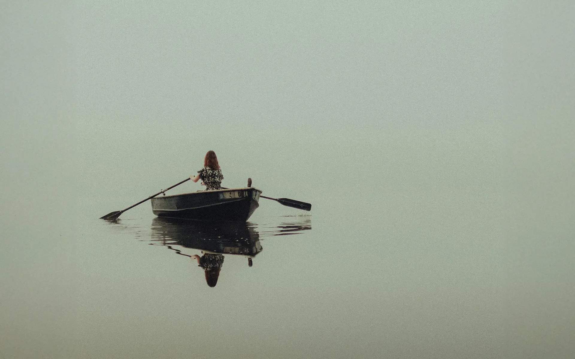 A woman with long hair sits in a small rowboat on a calm, misty body of water, paddling gently. The still water creates a near-perfect reflection of the boat and the woman. The muted, foggy atmosphere gives a sense of solitude and tranquility.