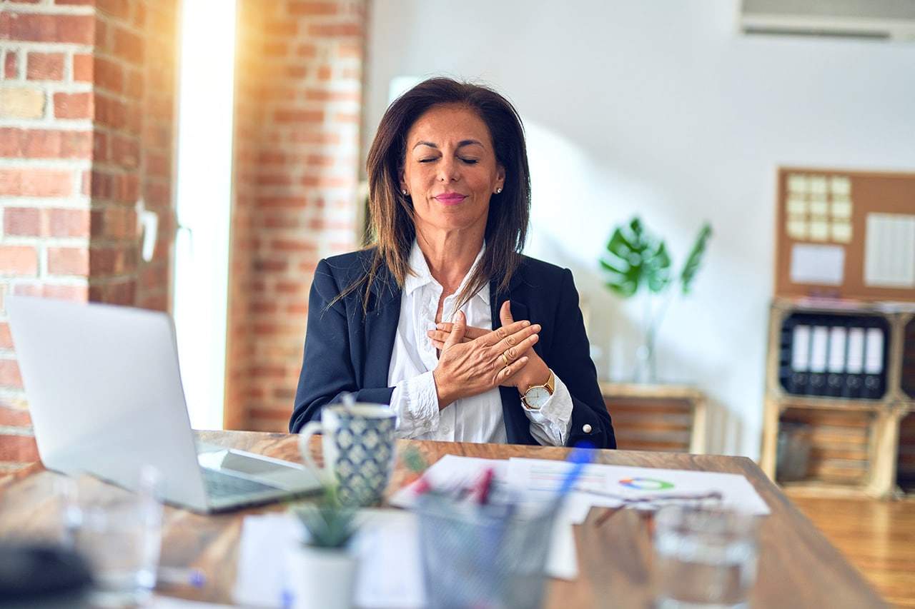 Woman at her desk at work, with eyes closed and hands on chest, giving herself self-compassion and radical self-care