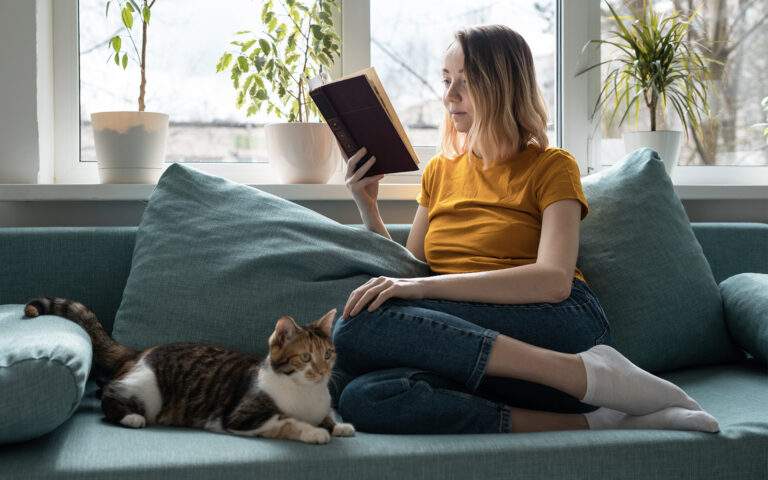 Photo of a woman curled up on a cozy couch by a window, reading a book, with her cat cuddling next to her.