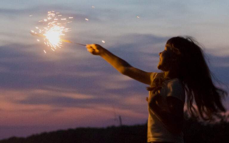 Photo of a girl standing outside at dusk, holding a lit sparkler aloft against the darkening sky