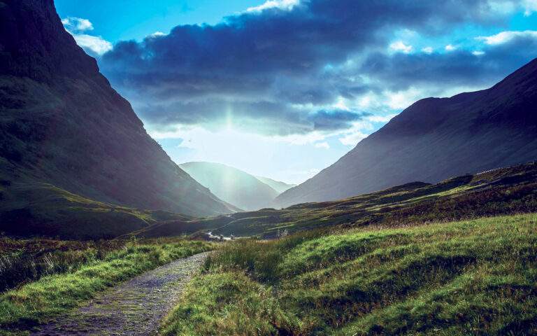 Photo of a beautiful path through a grassy valley with mountains and a sunny blue sky in the distance, suggesting positivity and good news.
