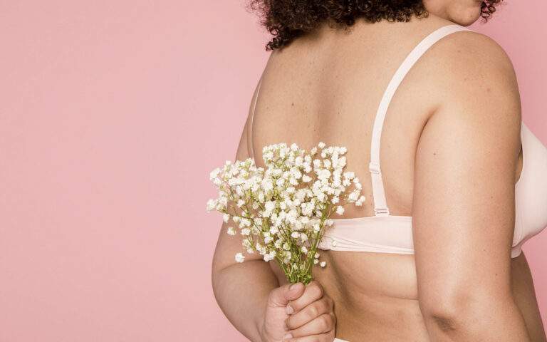 Photo from the back of a light skinned woman wearing a pink bra, holding a small bouquet of white flowers behind her back