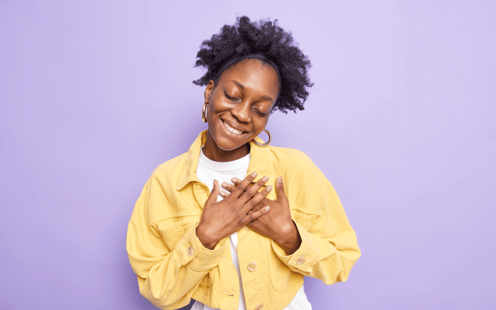 A young Black woman smiles and holds her hands over her heart in front of a purple studio background.
