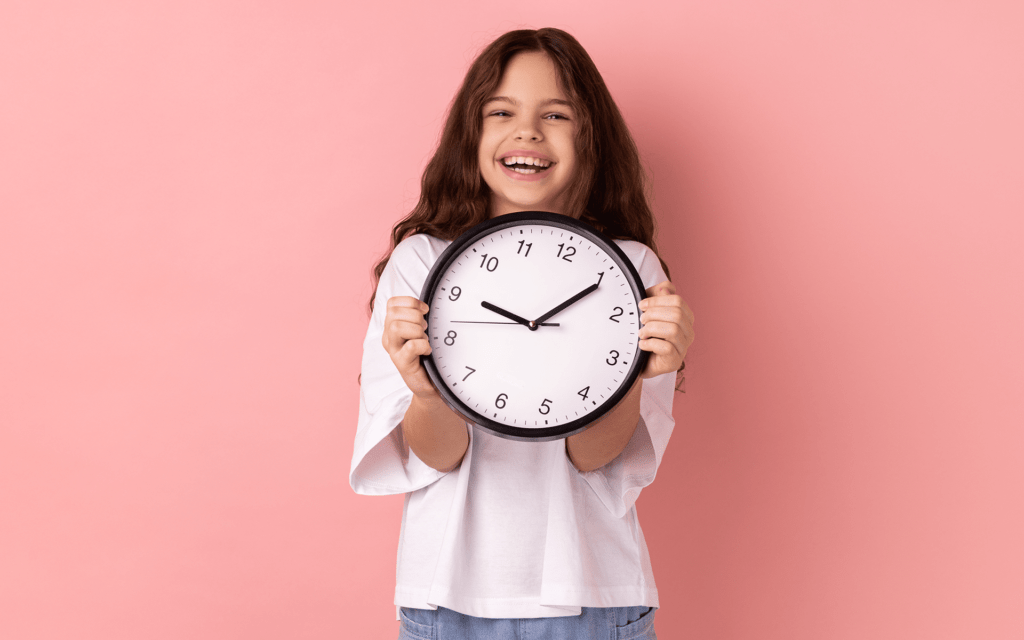 Photo of a young girl smiling and holding an analog clock while standing in front of a pink studio background.