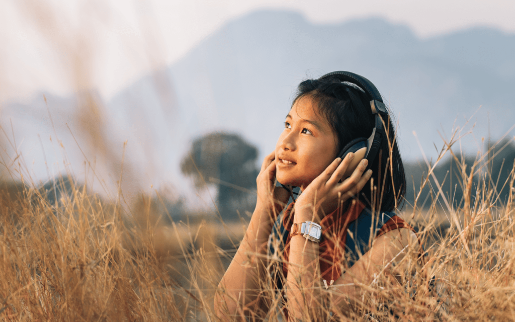 Photo of a young Asian girl in a field of tall grass smiling and wearing black headphones.