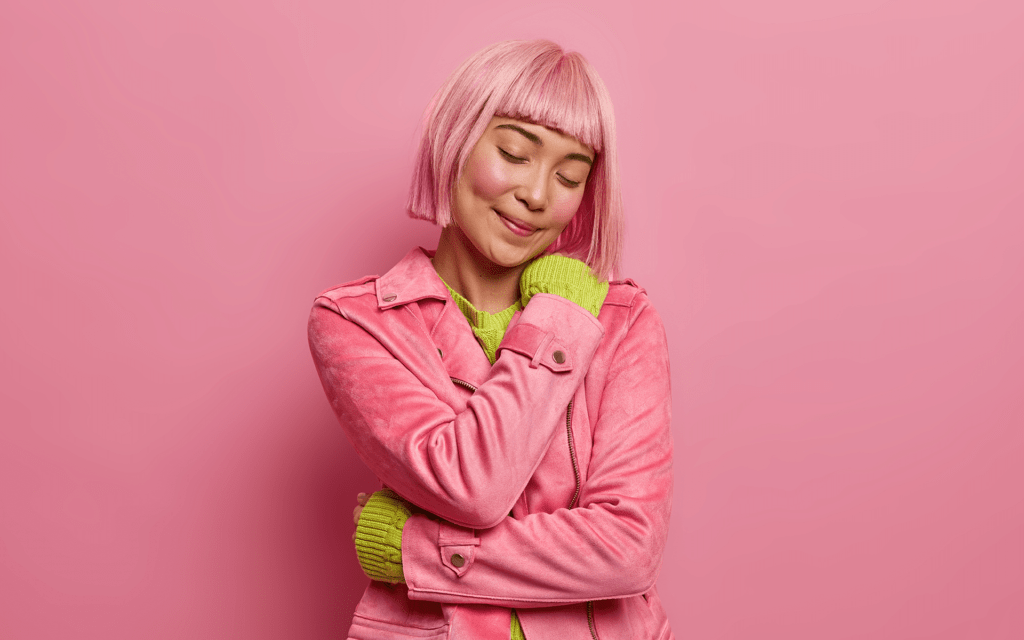 Photo of an Asian teenage girl giving herself a hug and smiling while standing in front of a pink studio background.